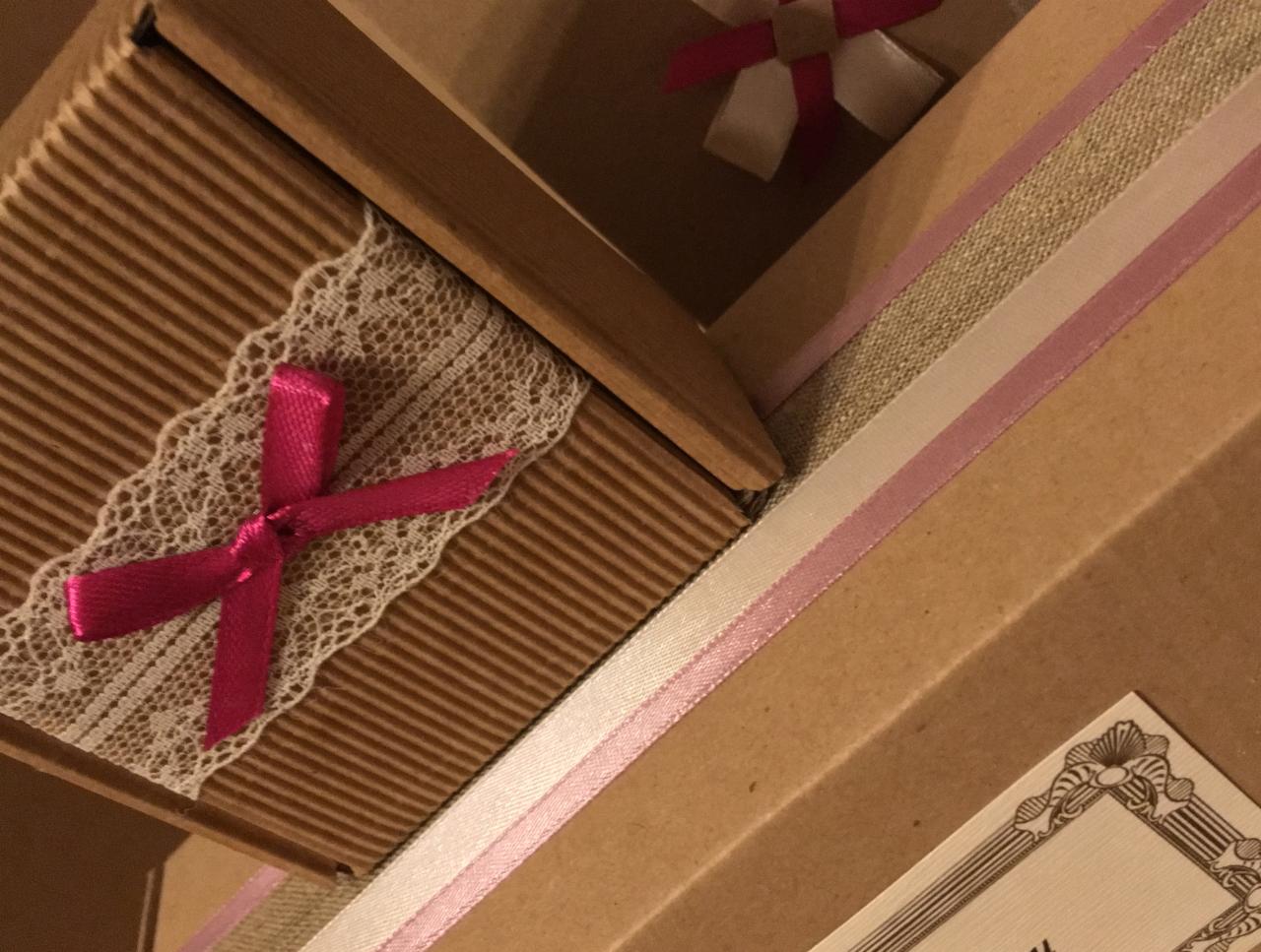 Brązowe pudełka z tektury falistej, ozdobione koronką, różowymi wstążkami i kokardkami, widoczne z góry pod kątem.