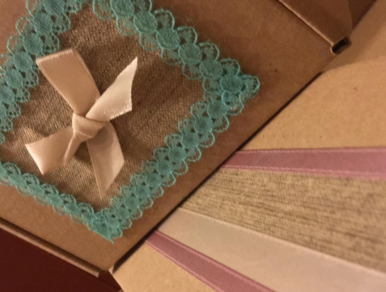 Ozdobne, tekturowe pudełko z jutowym kwadratem obramowanym turkusową koronką i beżową kokardką, widoczne fragmenty innych pudełek z ozdobnymi wstążkami.