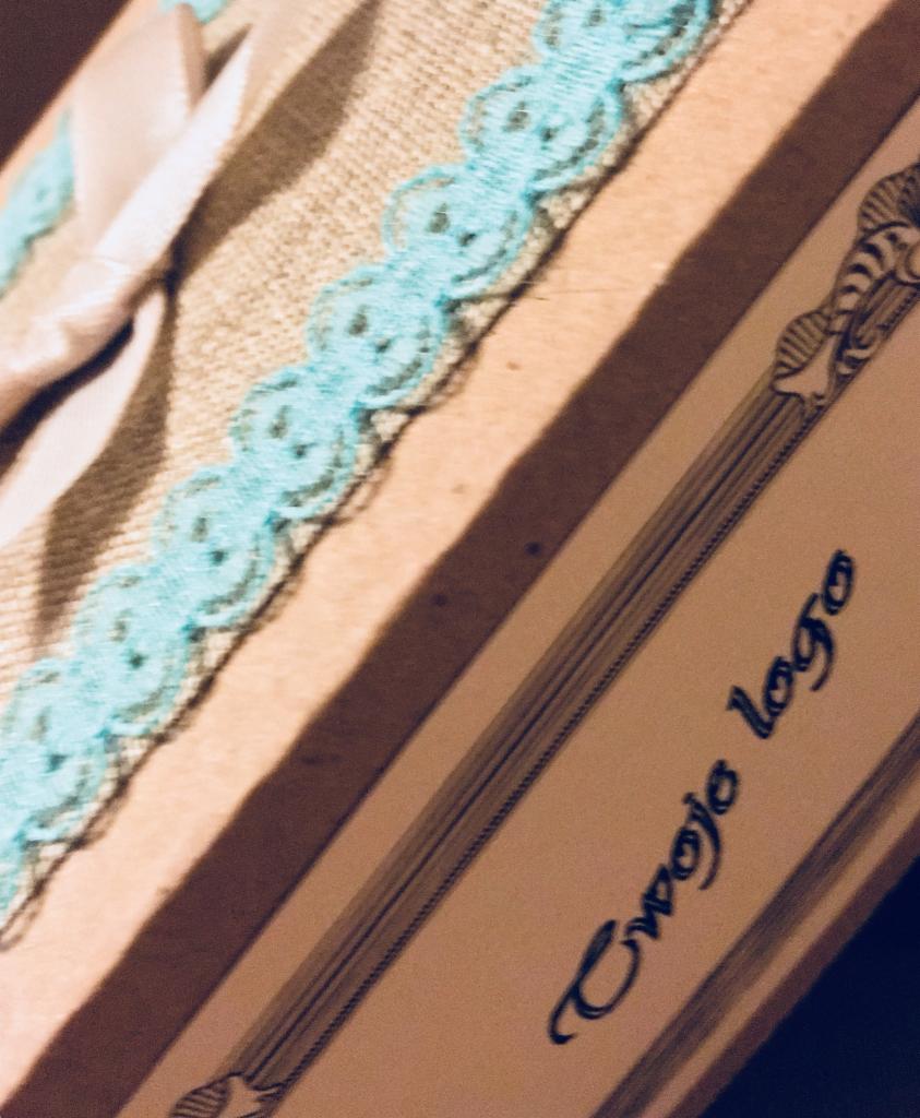 Beżowe pudełko z dekoracyjną, błękitną koronką, satynową wstążką i napisem 'Twoje logo' w ozdobnej czcionce.