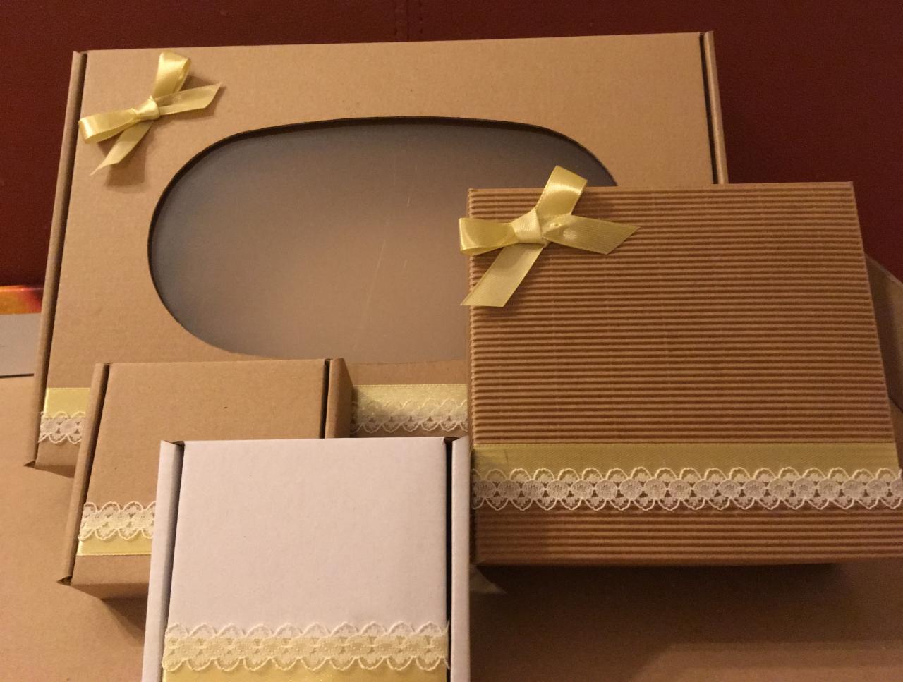 Zestaw ozdobnych pudełek z tektury falistej i gładkiej, przewiązanych wstążkami i koronką, w odcieniach brązu i bieli, ustawionych na brązowym tle.