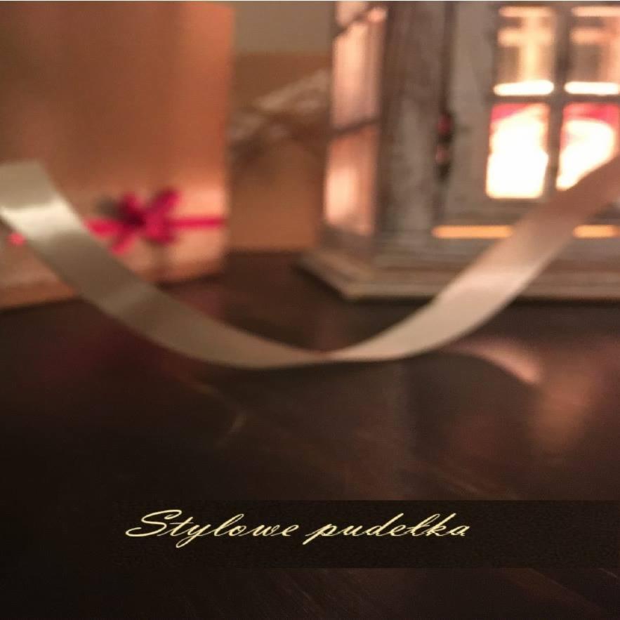 Ujęcie z bliska: wstążka satynowa opadająca na błyszczący stół, w tle pudełko prezentowe z czerwoną kokardą i dekoracyjna latarenka.