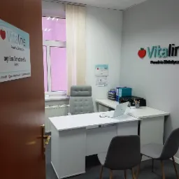 Wnętrze gabinetu poradni dietetycznej Vitaline z biurkiem, krzesłami dla pacjent&oacute;w i logo na ścianie. Na biurku laptop i przybory biurowe.