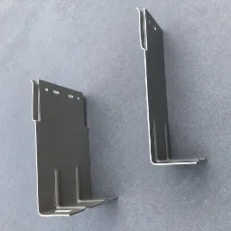 Dwa metalowe wsporniki elewacyjne w kształcie litery L, jeden z otworami, leżące na szarej powierzchni.