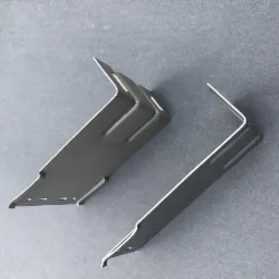 Dwa metalowe wsporniki kątowe na szarym tle, potencjalnie używane do mocowania elementów elewacji w Tczewie. Jeden wspornik posiada dodatkowe otwory i przetłoczenia.