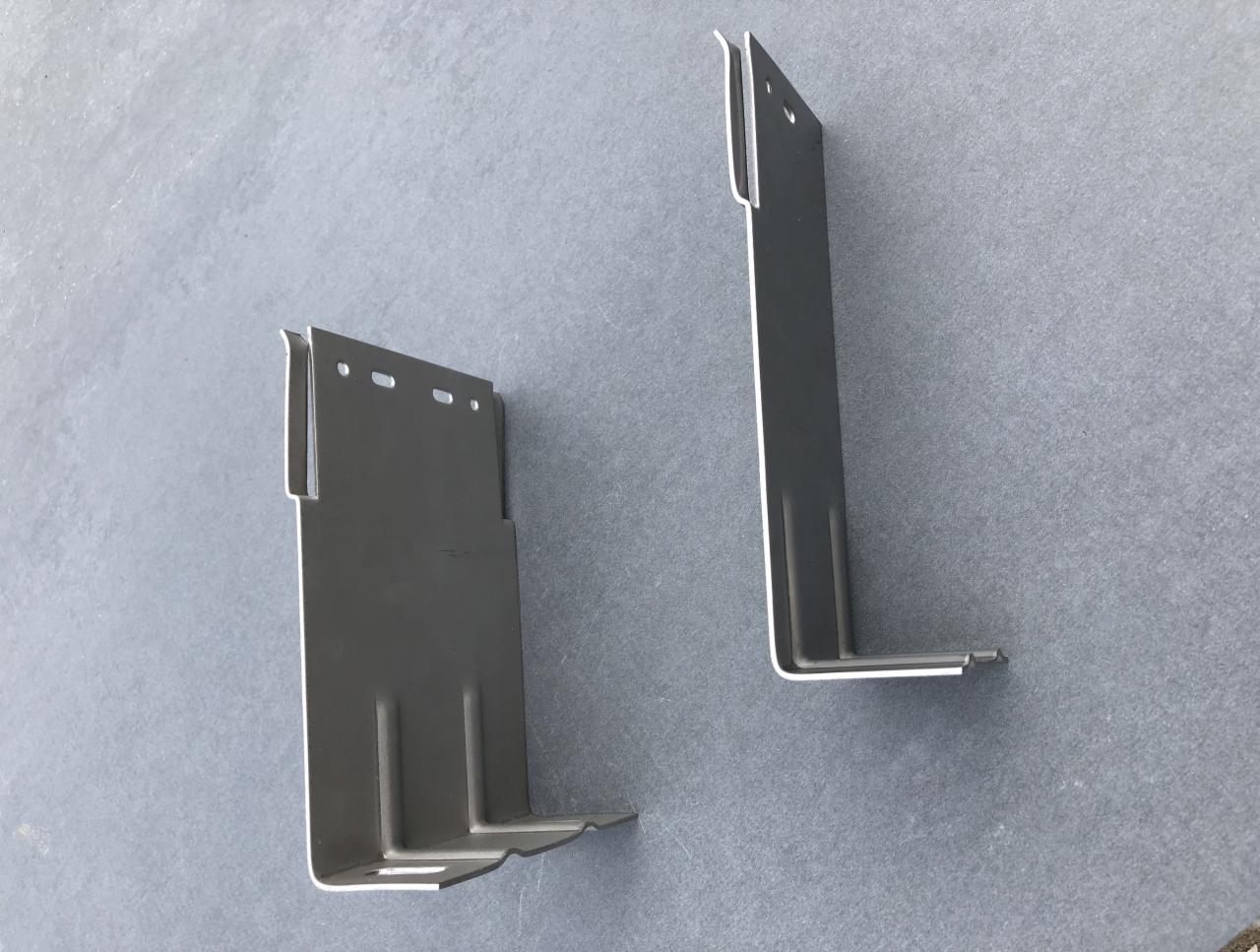 Dwa metalowe wsporniki elewacyjne w kształcie litery L, jeden z otworami, leżące na szarej powierzchni.