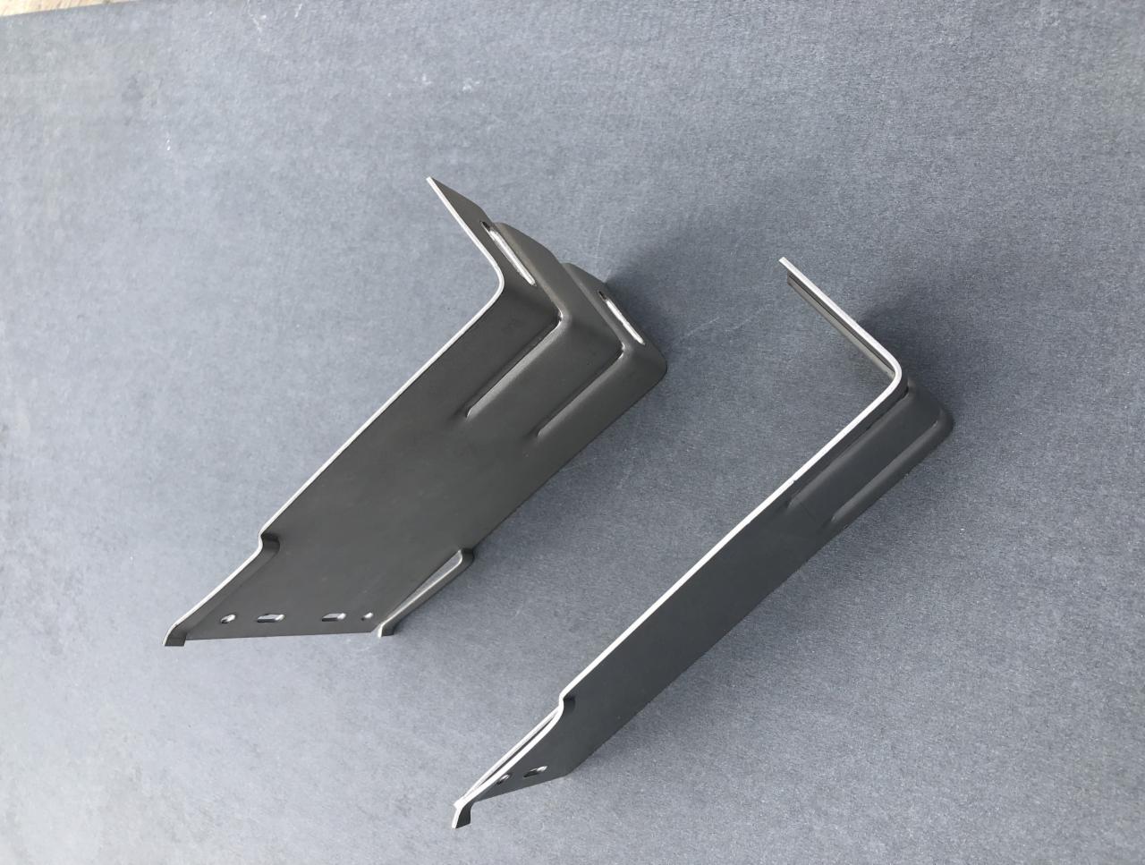 Dwa metalowe wsporniki kątowe na szarym tle, potencjalnie używane do mocowania elementów elewacji w Tczewie. Jeden wspornik posiada dodatkowe otwory i przetłoczenia.