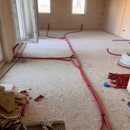 Woj-SAN - Wnętrze w stanie surowym z rozłożonymi czerwonymi rurami instalacji grzewczej podłogowej, narzędzia i materiały budowlane w rogu pomieszczenia, widoczne okna i drzwi balkonowe.