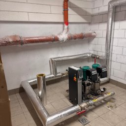 Woj-SAN - Kompletna instalacja pomp hydroforowych z zabezpieczonymi rurami w piwnicy, widoczne rury miedziane i plastikowe, izolacja termiczna oraz kratka ściekowa w podłodze.