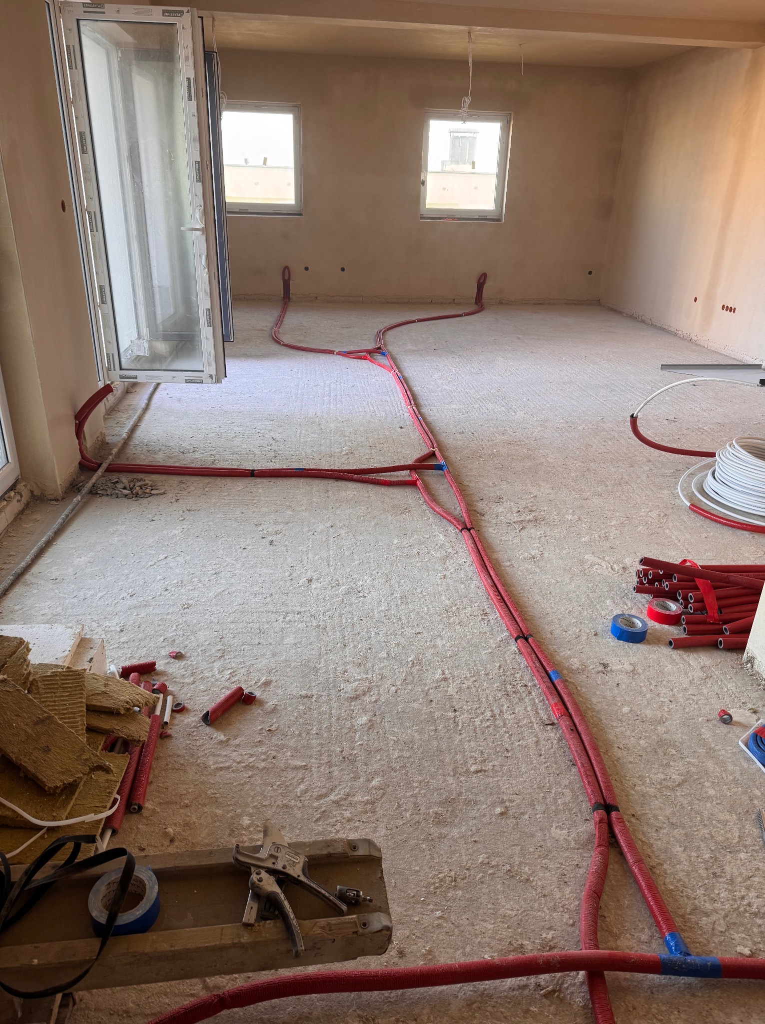 Wnętrze w stanie surowym z rozłożonymi czerwonymi rurami instalacji grzewczej podłogowej, narzędzia i materiały budowlane w rogu pomieszczenia, widoczne okna i drzwi balkonowe.