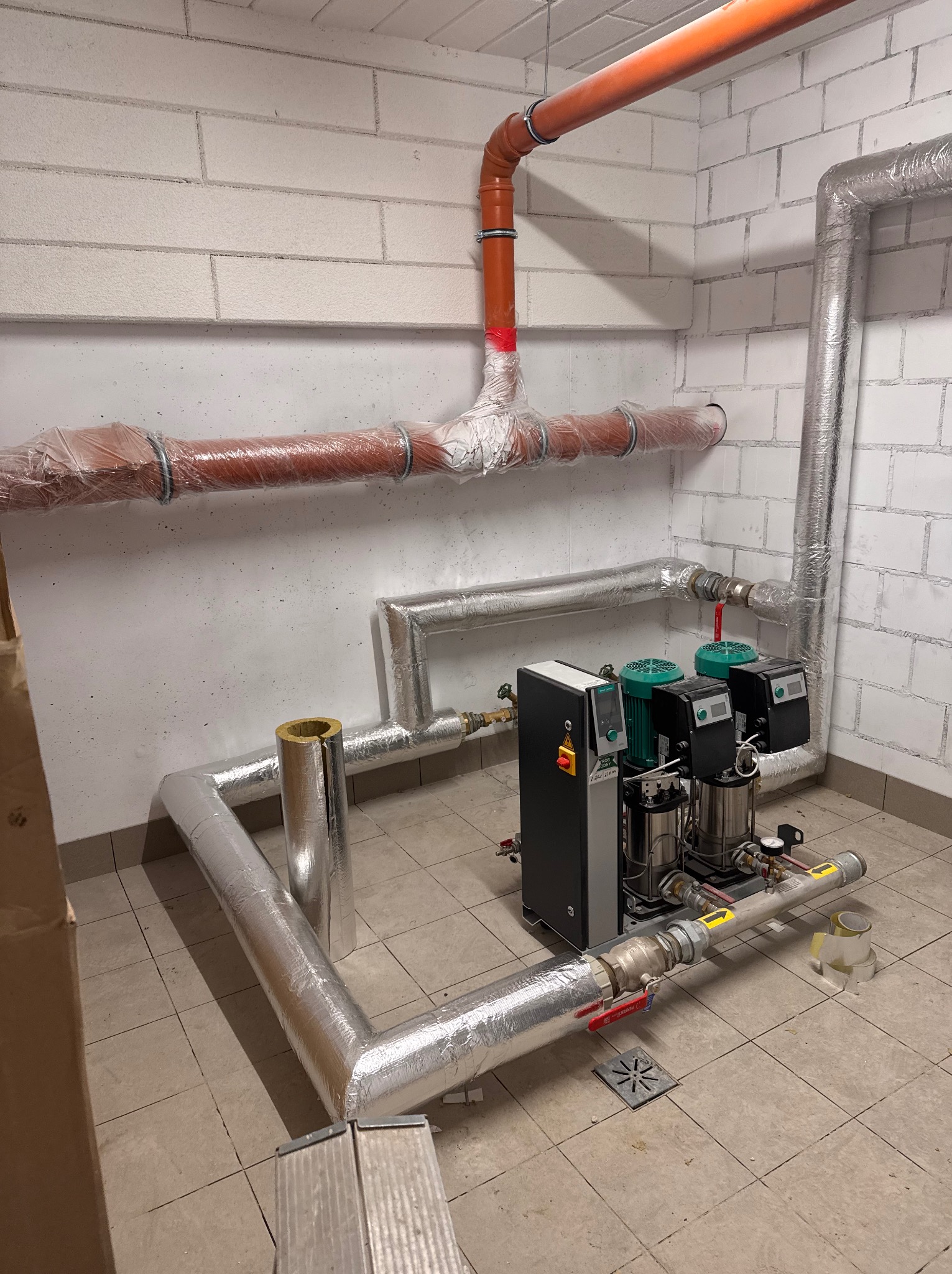 Kompletna instalacja pomp hydroforowych z zabezpieczonymi rurami w piwnicy, widoczne rury miedziane i plastikowe, izolacja termiczna oraz kratka ściekowa w podłodze.