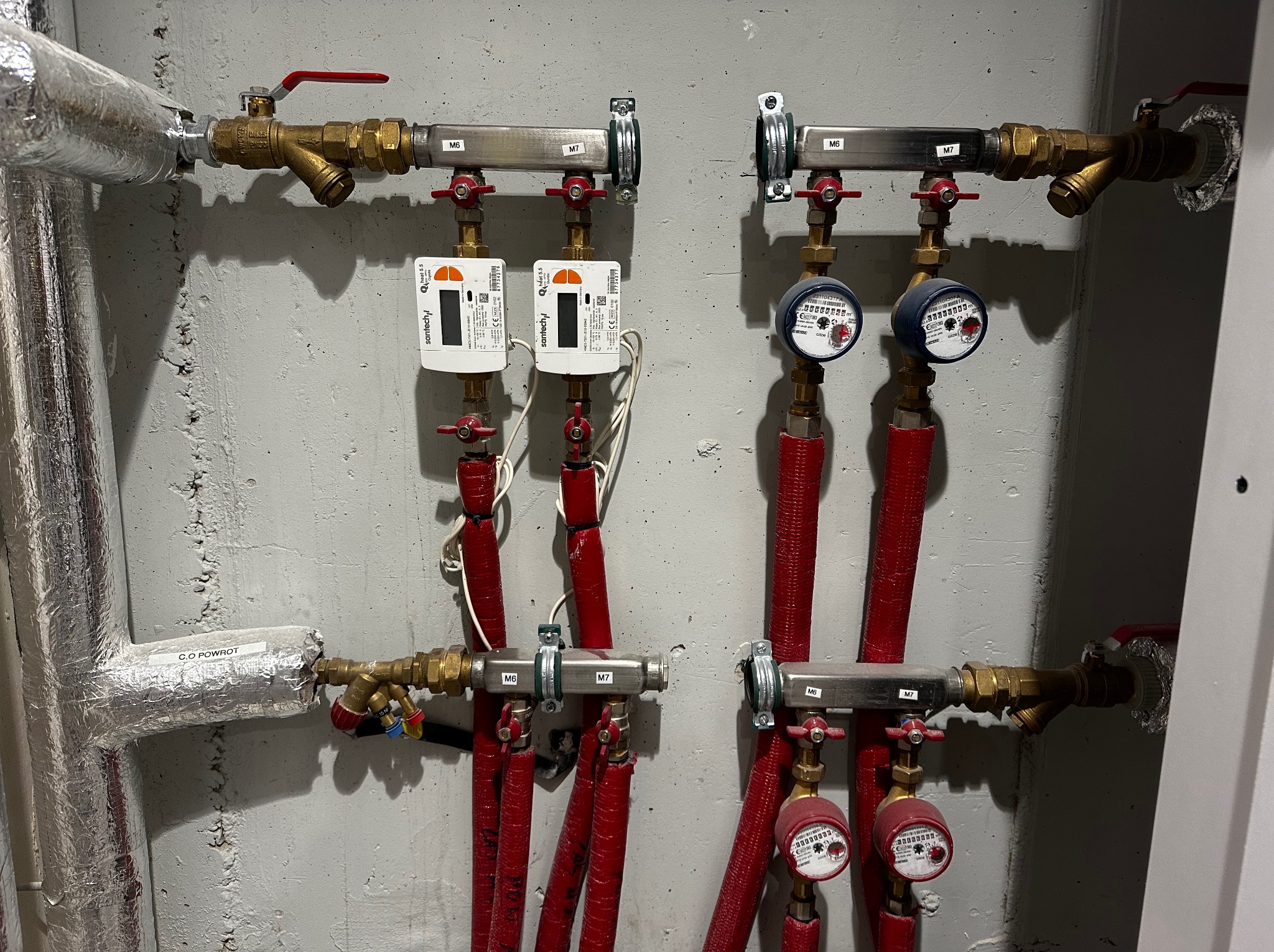 Rozdzielacz instalacji grzewczej z licznikami ciepła i zaworami, rury w czerwonej izolacji, etykiety M6 i M7 na elementach metalowych, widoczne przyłącze powrotu C.O. z izolacją.
