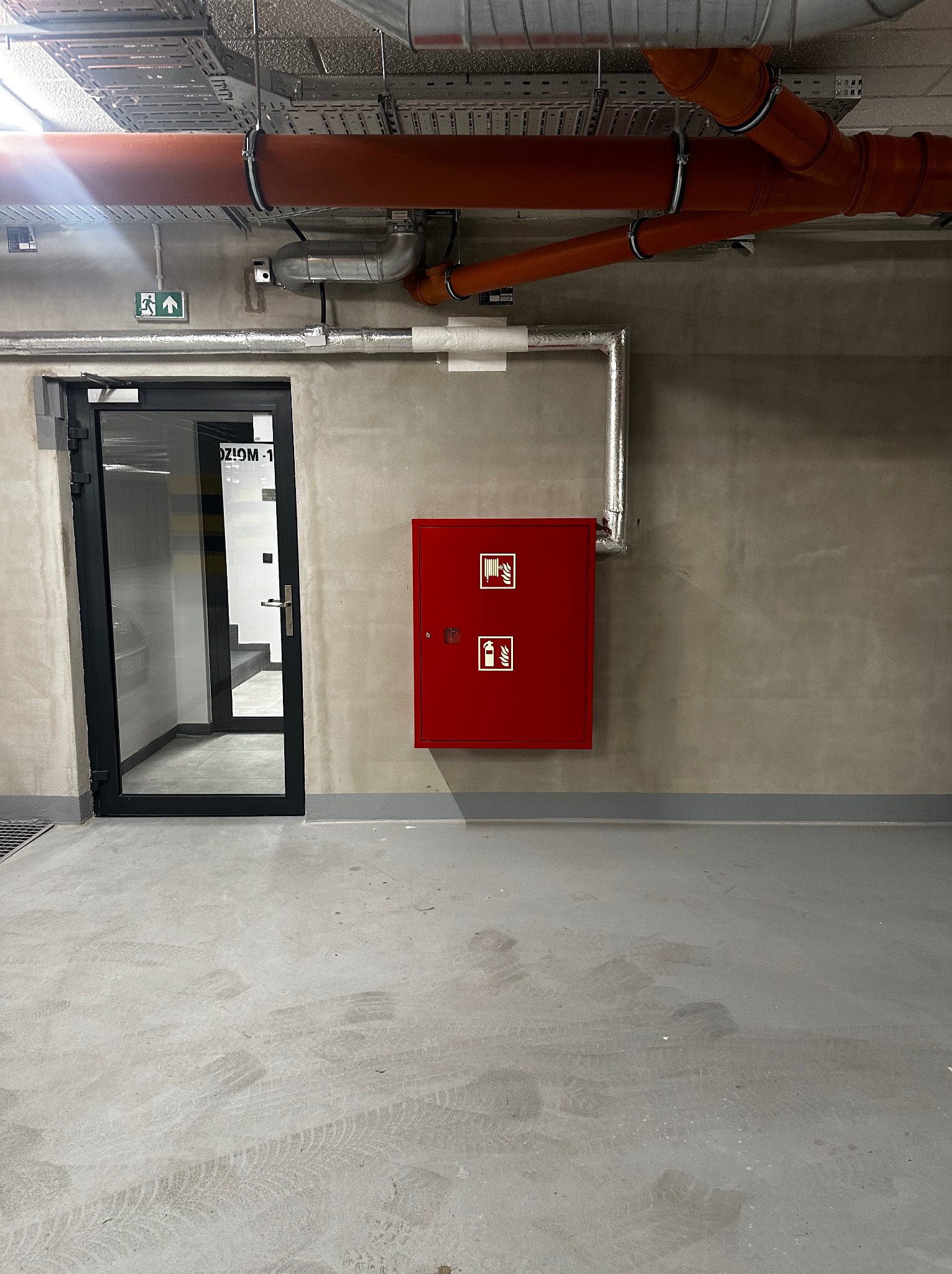 Wnętrze garażu podziemnego z widocznymi rurami instalacji wentylacyjnej pod sufitem, czerwoną szafką hydrantową na ścianie oraz wyjściem ewakuacyjnym z lewej strony.
