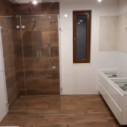 Nowoczesna łazienka w trakcie wykończenia z kabiną prysznicową typu walk-in z panelami imitującymi drewno, białymi płytkami na ścianach i częściowo zamontowaną białą szafką pod umywalkę.