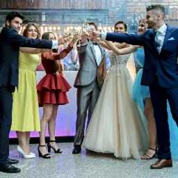 Grupa elegancko ubranych osób wznosi toast szampanem podczas uroczystości, z widocznym oświetleniem i dekoracjami w tle.