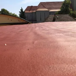 Czerwona, gumowa powierzchnia dachu z widocznymi drobnymi zanieczyszczeniami, w tle budynek z szarymi ścianami i czerwoną dachówką oraz wieża zegarowa.