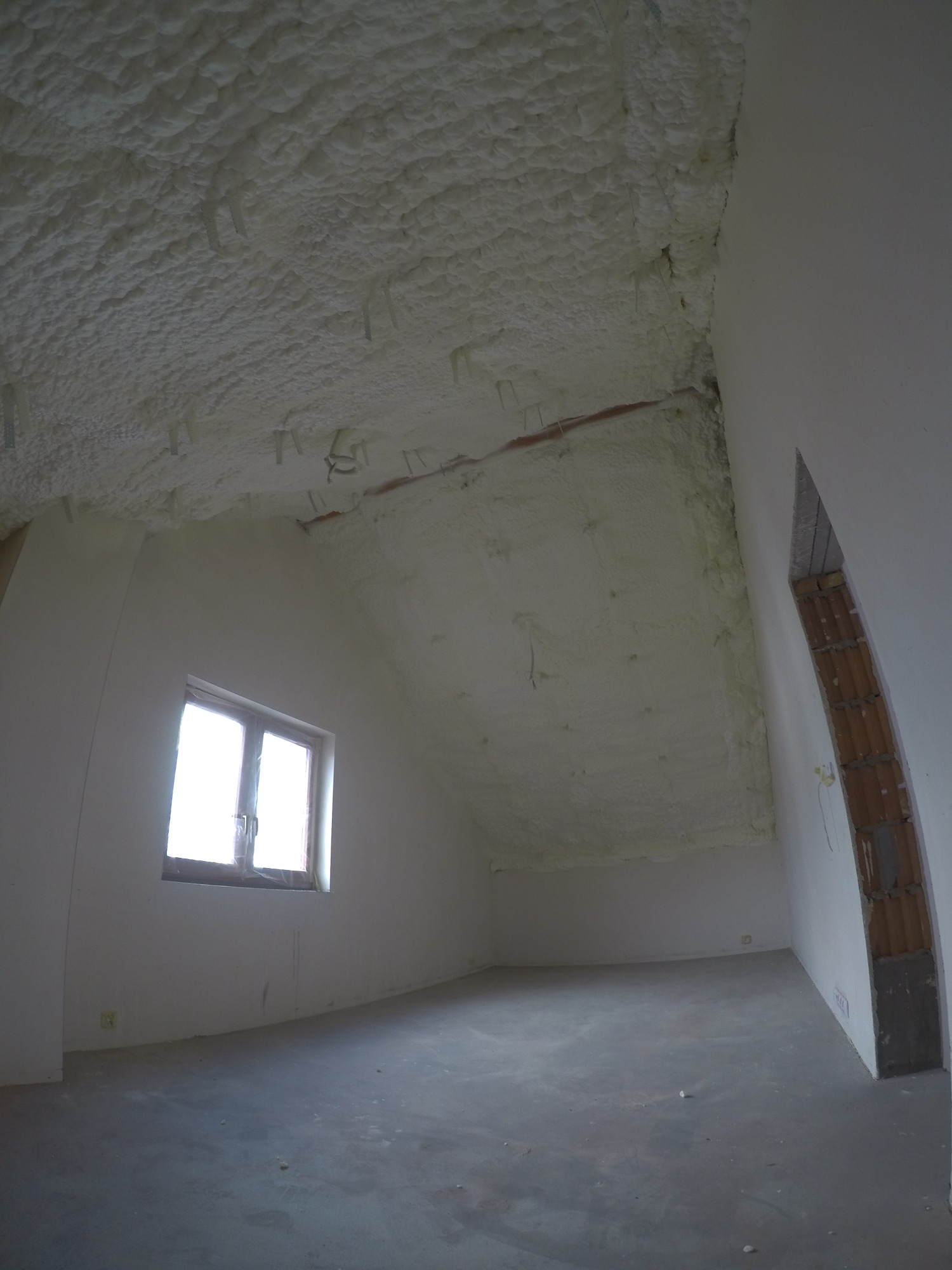 Poddasze po ociepleniu pianką PUR, widoczne nierównomierne pokrycie pianką i wystające elementy montażowe, okno w ścianie szczytowej, surowa podłoga.