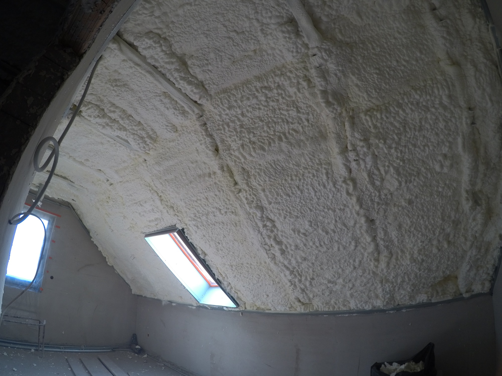 Poddasze po ociepleniu pianką PUR, widoczne okno dachowe i fragment instalacji elektrycznej, nierównomierna struktura piany.