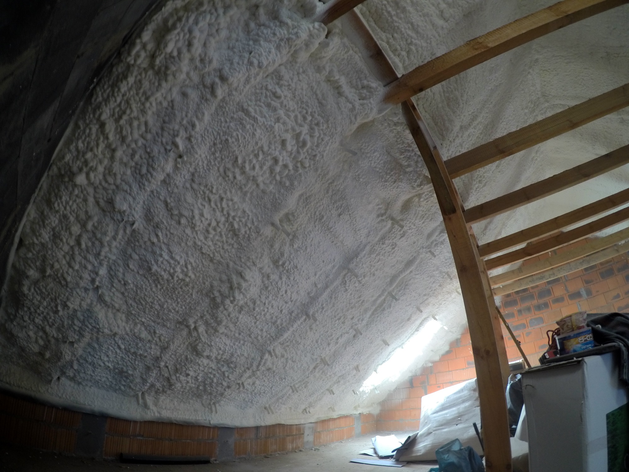Poddasze ocieplone białą pianką poliuretanową, widoczne krokwie drewniane, ściana z cegły, fragment okna dachowego i przedmioty w pomieszczeniu.