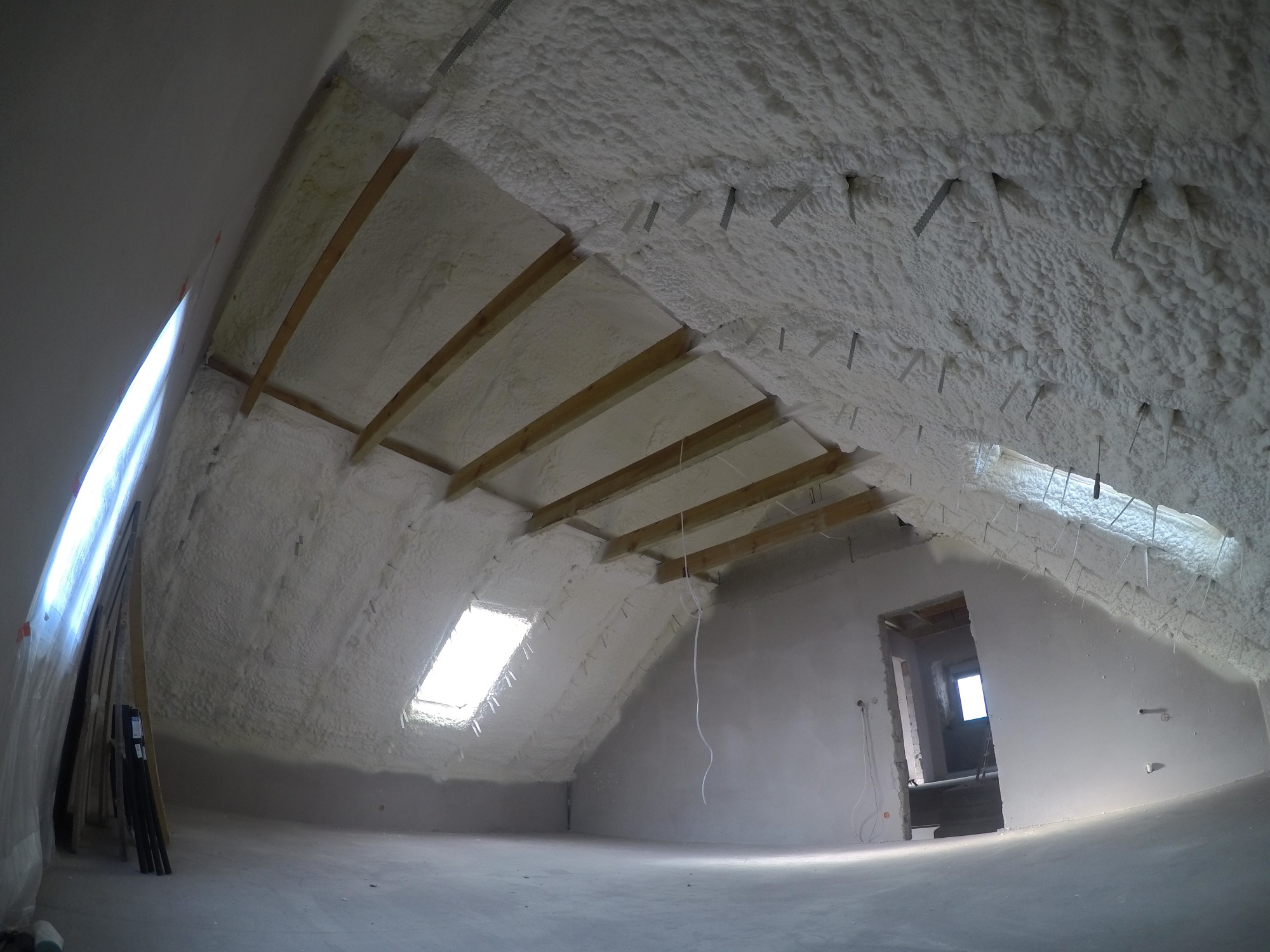 Poddasze po ociepleniu pianką: widoczne drewniane belki konstrukcyjne i warstwa izolacji z pianki PUR, okno dachowe w skosie, surowy stan wykończenia ścian i podłogi.