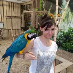 Młoda kobieta z upiętymi włosami trzyma na przedramieniu dużą, kolorową arę w pomieszczeniu przypominającym wolierę.