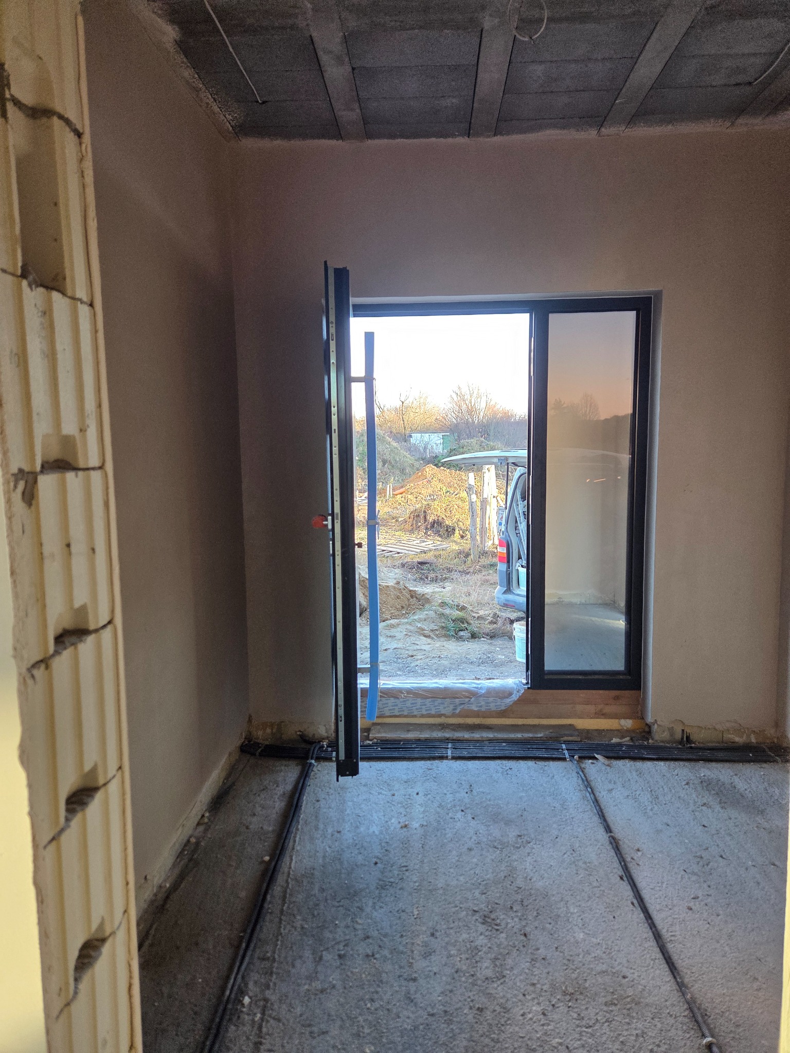 Wnętrze w stanie surowym: ściany po nałożeniu tynku, widoczne przewody na podłodze i otwarte drzwi na zewnątrz. Surowy betonowy strop.