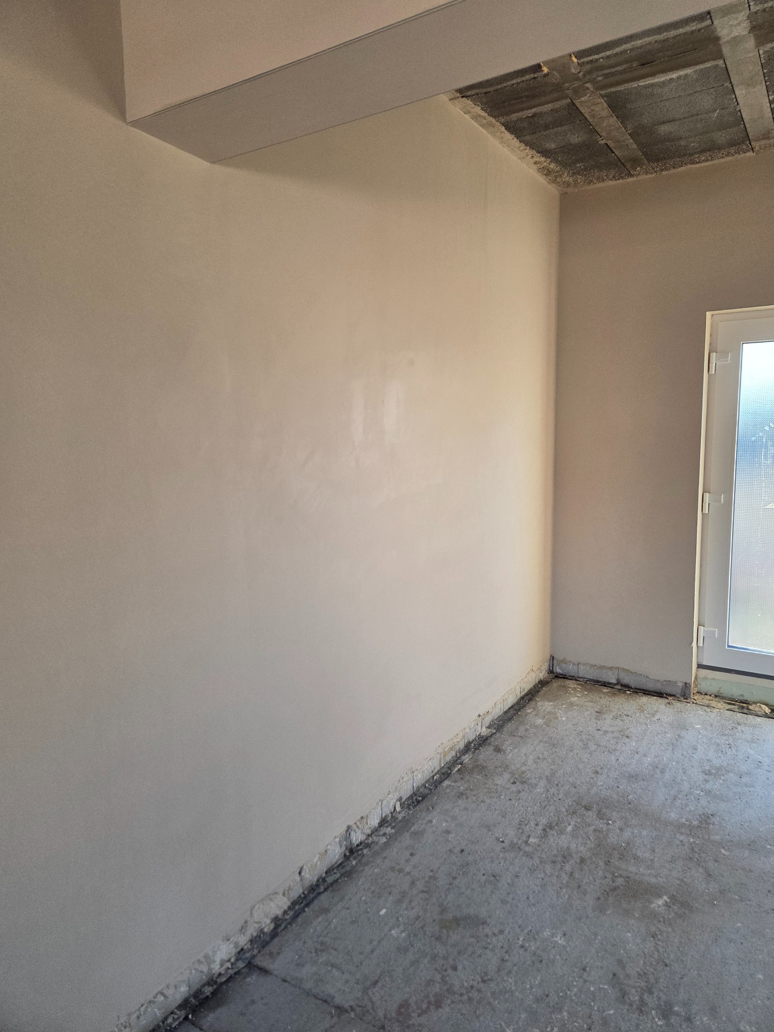Gładkie, jasne tynki na ścianach w rogu pokoju, surowy betonowy strop, widoczne ślady po pracach wykończeniowych na podłodze, białe drzwi z szybą w tle.