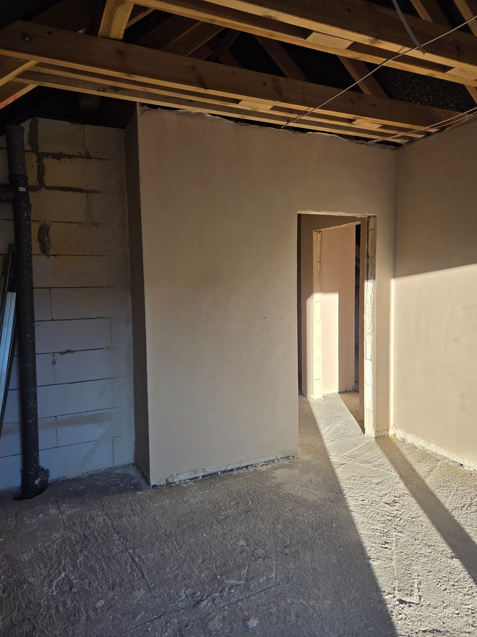 Wnętrze w trakcie prac wykończeniowych: ściany pokryte świeżym tynkiem, widoczne surowe drewniane belki stropowe i fragment ściany z bloczków. Wpadające światło słoneczne.