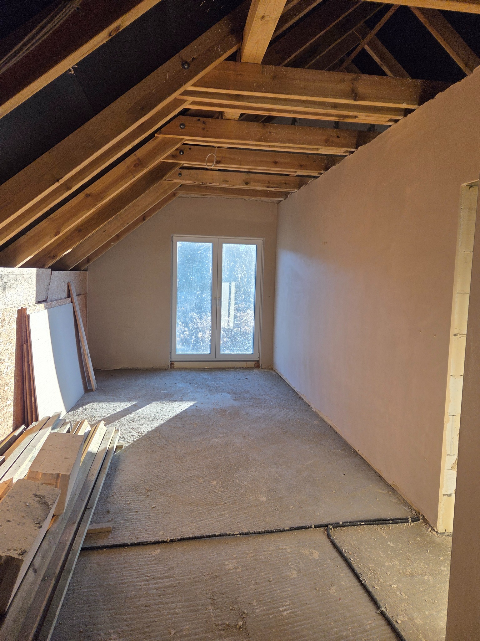 Poddasze po nałożeniu tynku maszynowego na ścianach, widoczne drewniane elementy konstrukcji dachu i okno. Na podłodze leżą materiały budowlane.