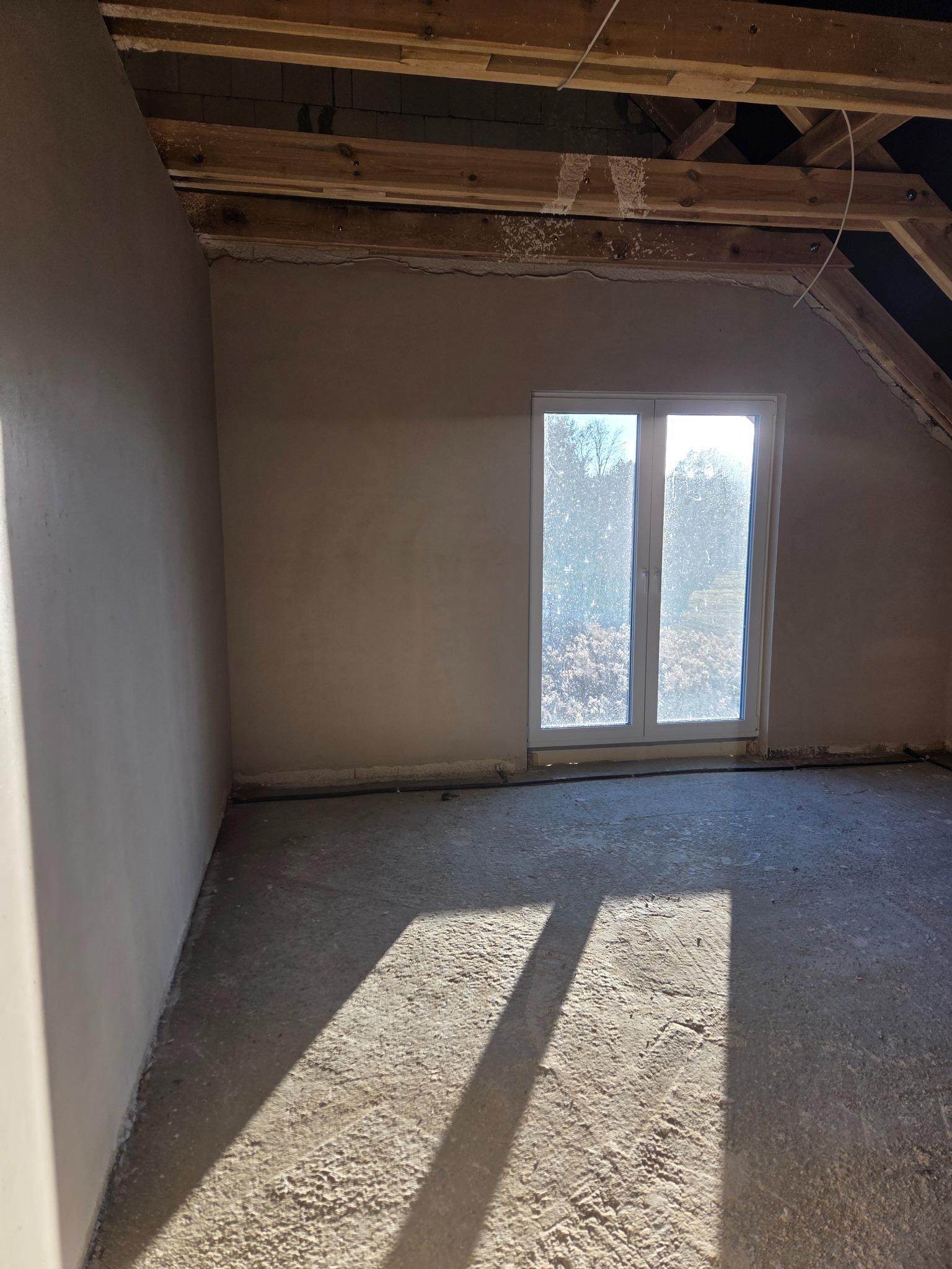 Pokój na poddaszu po tynkowaniu maszynowym ścian, widoczne drewniane elementy konstrukcji dachu i okno z widokiem na zewnątrz. Świeże tynki w jasnym odcieniu.