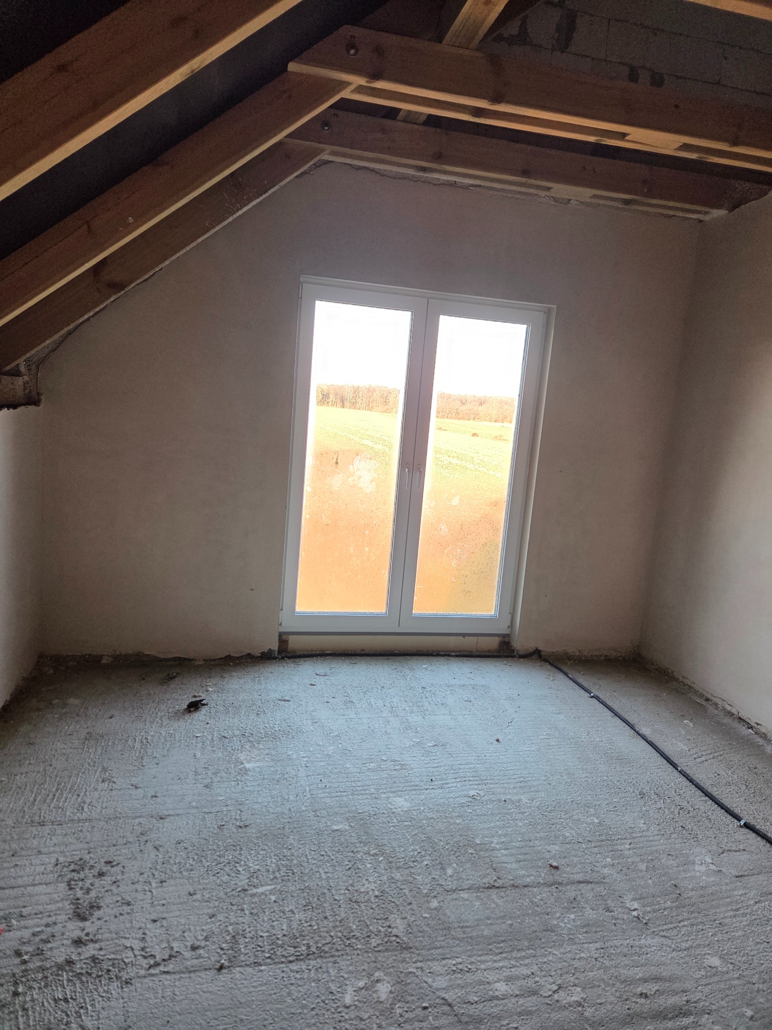 Poddasze po nałożeniu tynków: gładkie ściany w odcieniach beżu, białe okno z widokiem na pola, surowy beton na podłodze i widoczne elementy drewnianej konstrukcji dachu.
