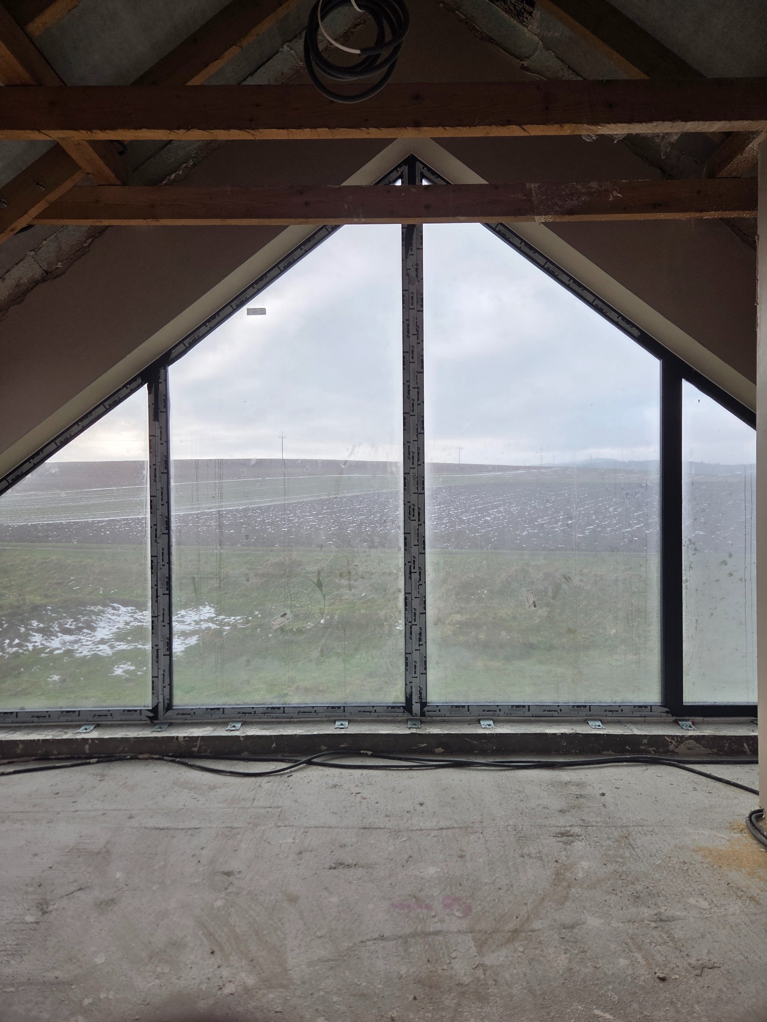 Poddasze z oknem szprosowym w kształcie trapezu. Surowy tynk na ścianach, betonowa podłoga. Widok na zaśnieżone pole za oknem. Prace wykończeniowe.