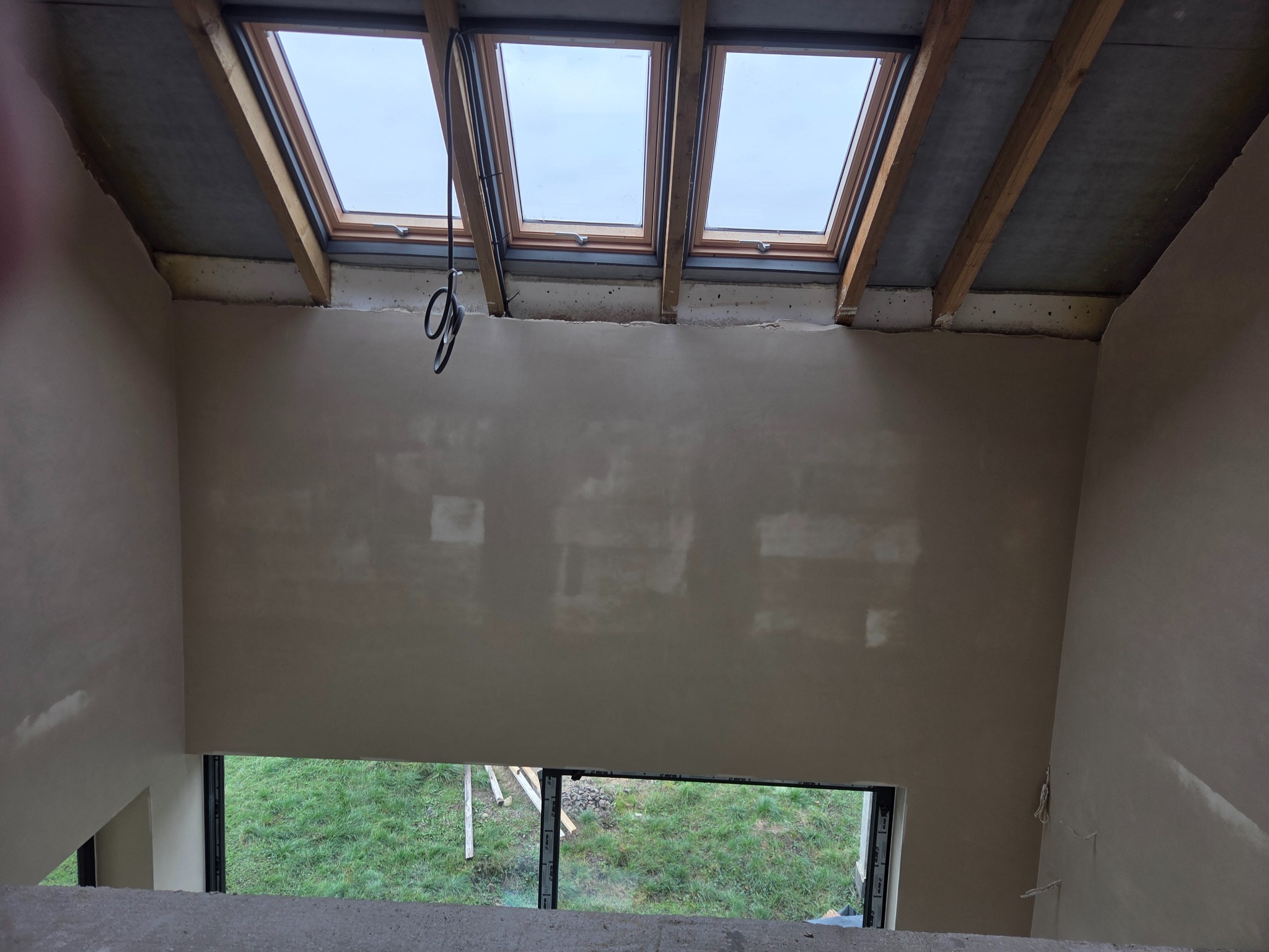 Wnętrze po nałożeniu tynków: ściany o gładkiej fakturze, okna dachowe z drewnianą ramą, widok na zewnątrz przez okno. Kabel zwisa z sufitu.