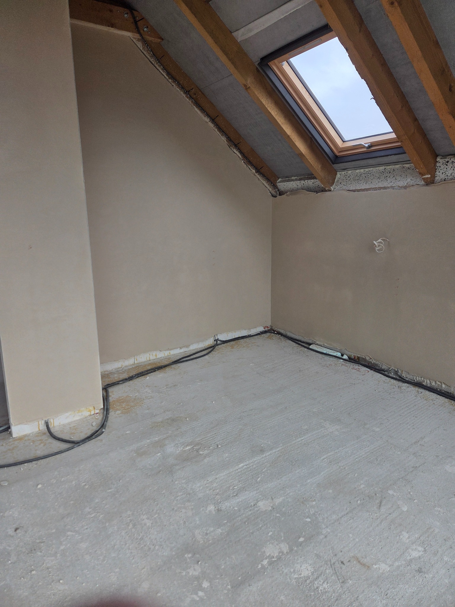 Wnętrze poddasza po nałożeniu tynków, widoczne okno dachowe i kable przy podłodze. Ściany gładkie, przygotowane do dalszych prac wykończeniowych.