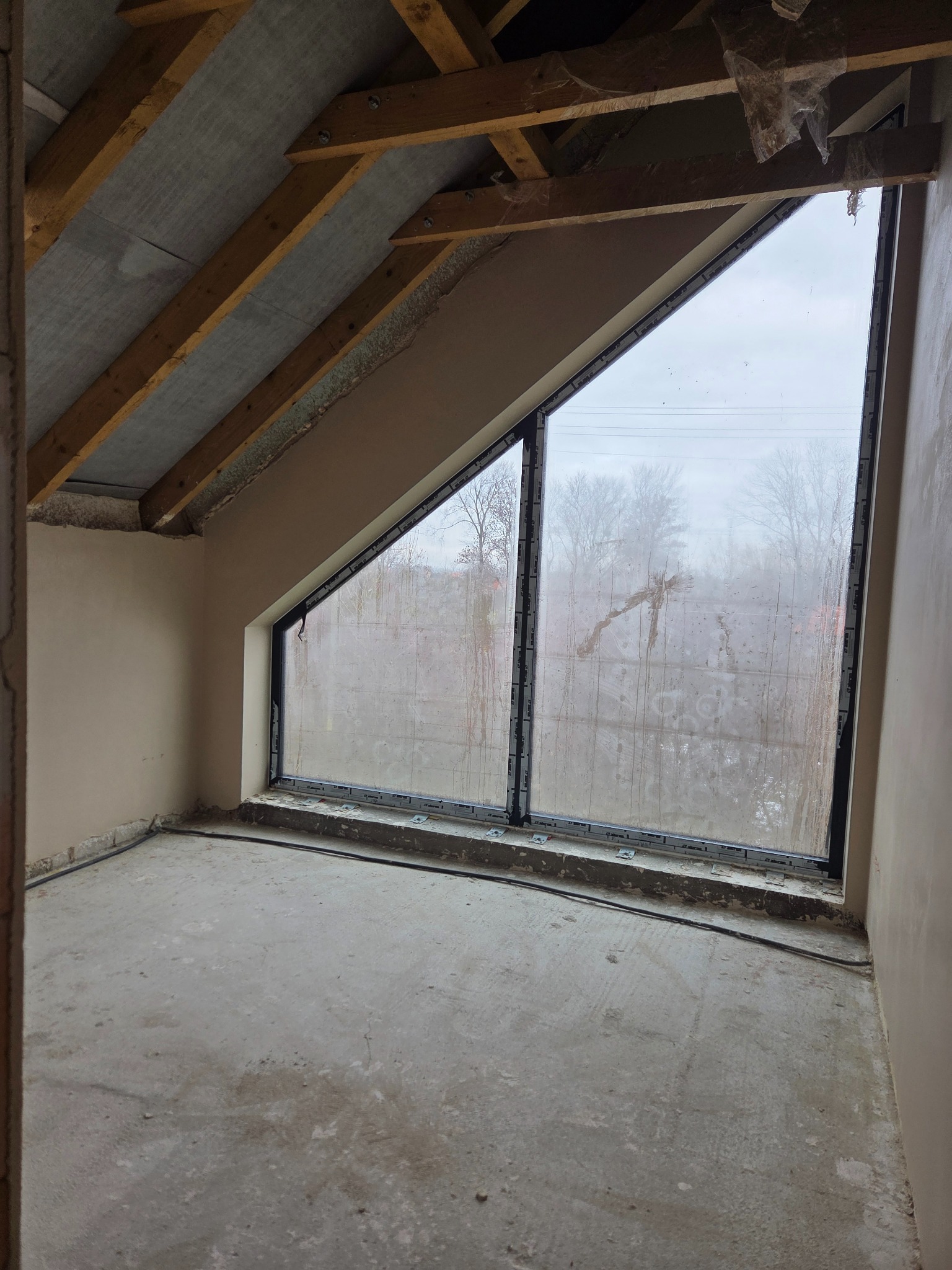 Surowe wnętrze poddasza z oknem o nietypowym kształcie, ściany przygotowane do tynkowania, widoczne drewniane elementy konstrukcji dachu. Stan surowy.
