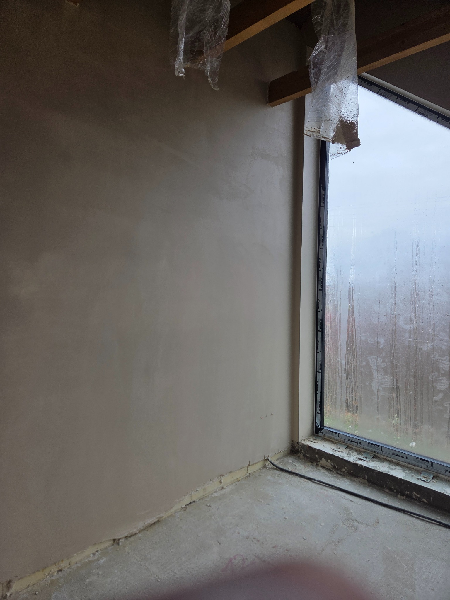 Gładka ściana po tynkowaniu maszynowym w Strzelinie, widok z rogu pomieszczenia, okno z zaparowaną szybą. Folia ochronna na elementach konstrukcyjnych.