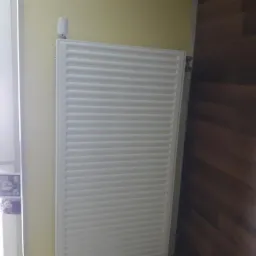 Biały, panelowy grzejnik z termostatem, zamontowany na żółtej ścianie, obok okna i drewnianej podłogi.