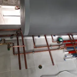 Szare zasobnik ciepłej wody z miedzianą instalacją hydrauliczną, widoczne rury w izolacji czerwonej i niebieskiej, manometr i elementy montażowe na tle szarej płytki ceramicznej.