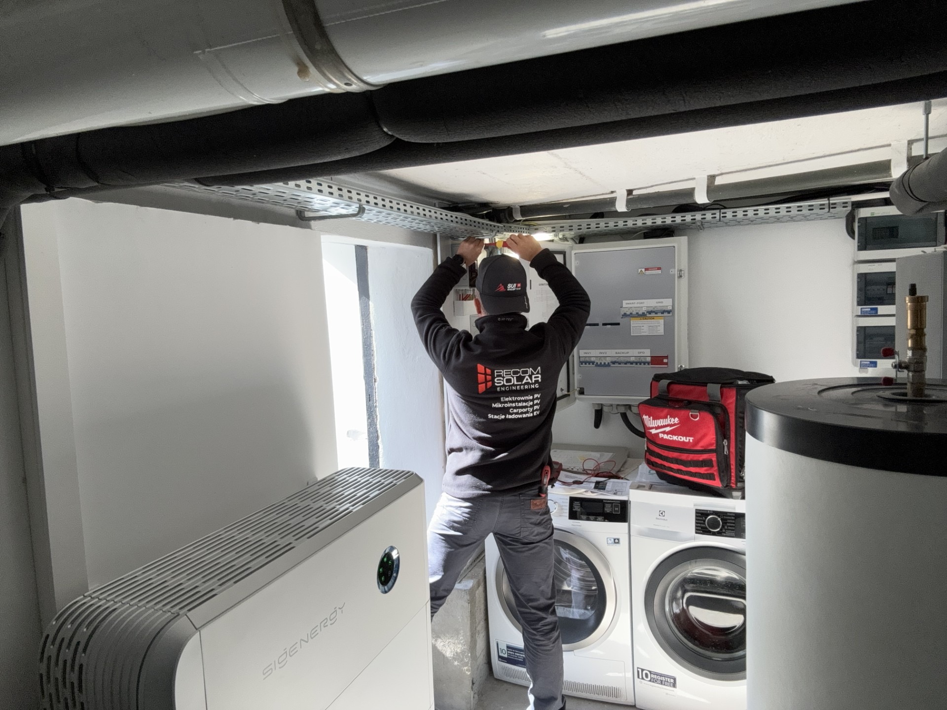 Instalator w trakcie montażu instalacji fotowoltaicznej w pomieszczeniu gospodarczym, widoczny inwerter, rozdzielnica, zasobnik ciepłej wody i pralka.
