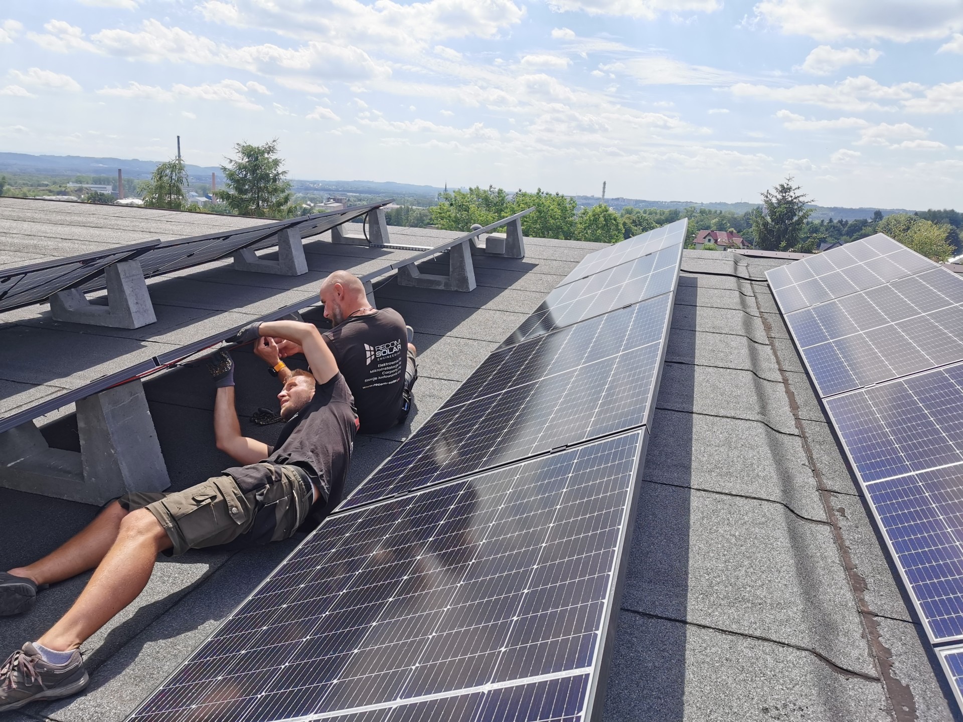 Montaż paneli fotowoltaicznych na dachu z widokiem na okolicę. Dwóch instalatorów w trakcie pracy, jeden leżący, drugi siedzący. Widoczne panele i elementy montażowe.