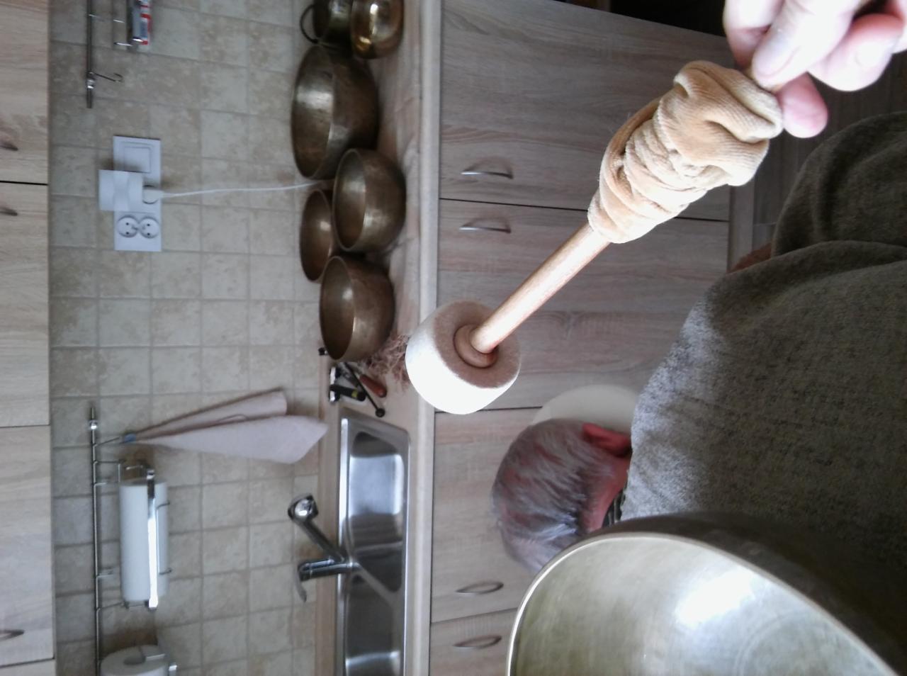 Osoba uderza pałeczką w misę dźwiękową, w tle widoczne inne misy i wyposażenie kuchni.