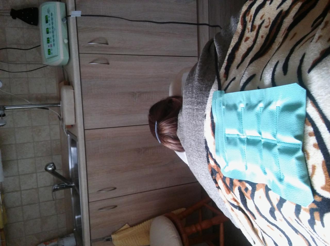 Pacjentka leży na leżance z poduszką chłodzącą na plecach, widoczna część głowy, w tle szafki i umywalka w gabinecie.