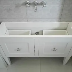 Biała, lakierowana szafka pod umywalkę z dwiema szufladami i srebrnymi uchwytami, bez umywalki, na tle jasnych płytek z delikatnym wzorem.