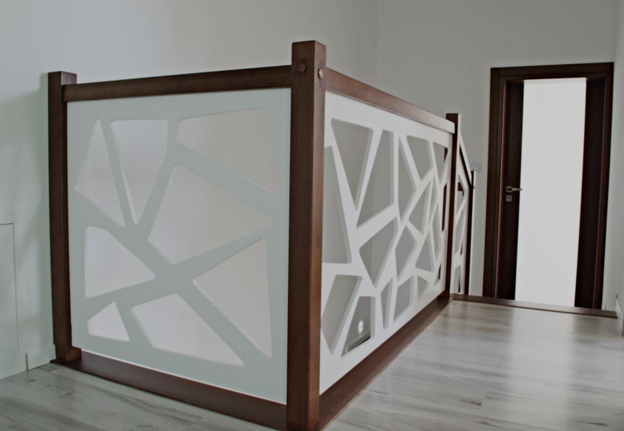 Barierka antresoli z drewnianą ramą i białym wypełnieniem o geometrycznym wzorze, widoczna na tle białych ścian i szarej podłogi, z ciemnymi drzwiami w tle.