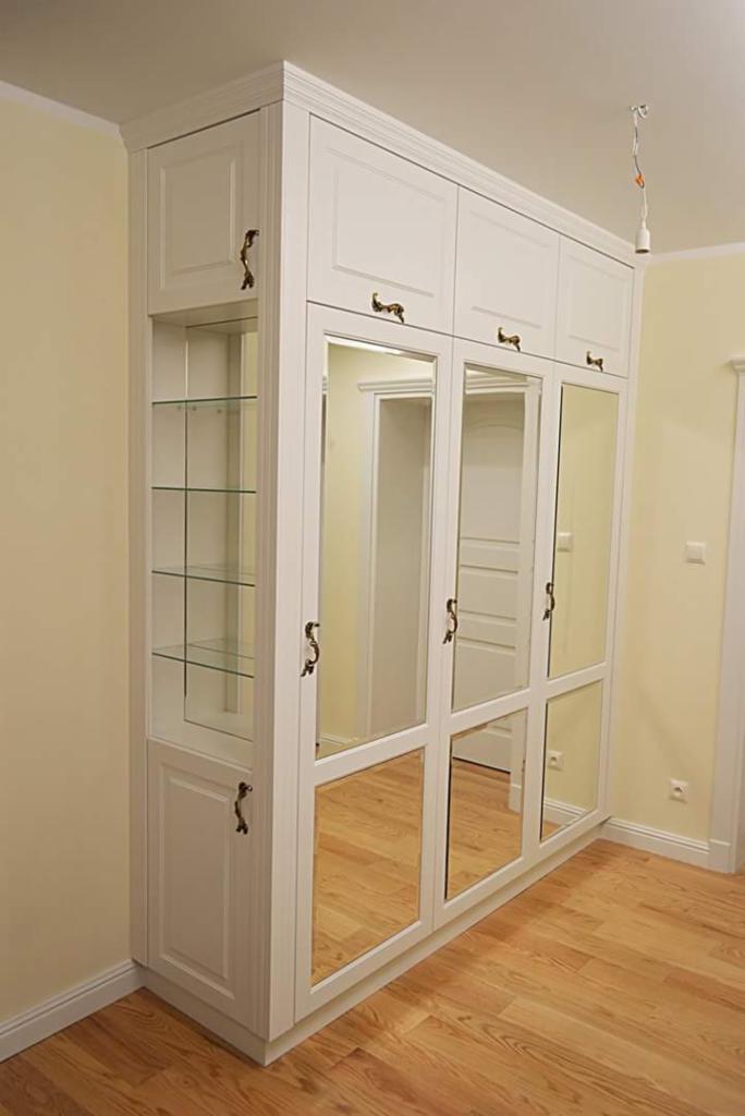 Biała szafa wnękowa z lustrami i przeszkloną witryną, drewniana podłoga, widoczne mosiężne uchwyty.