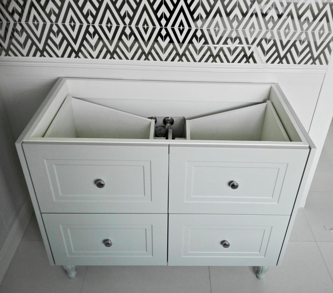 Biała szafka pod umywalkę z czterema szufladami, widoczne wnętrze bez umywalki i armatury, na tle biało-czarnej wzorzystej ściany.