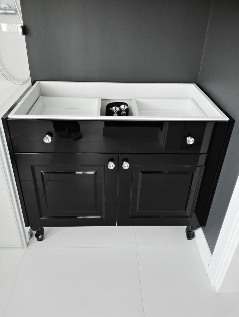 Czarna szafka pod umywalkę z połyskiem, z dekoracyjnymi kryształowymi uchwytami, na niskich nóżkach, z otwartą przestrzenią na umywalkę i armaturę, ustawiona na białych płytkach w łazience.