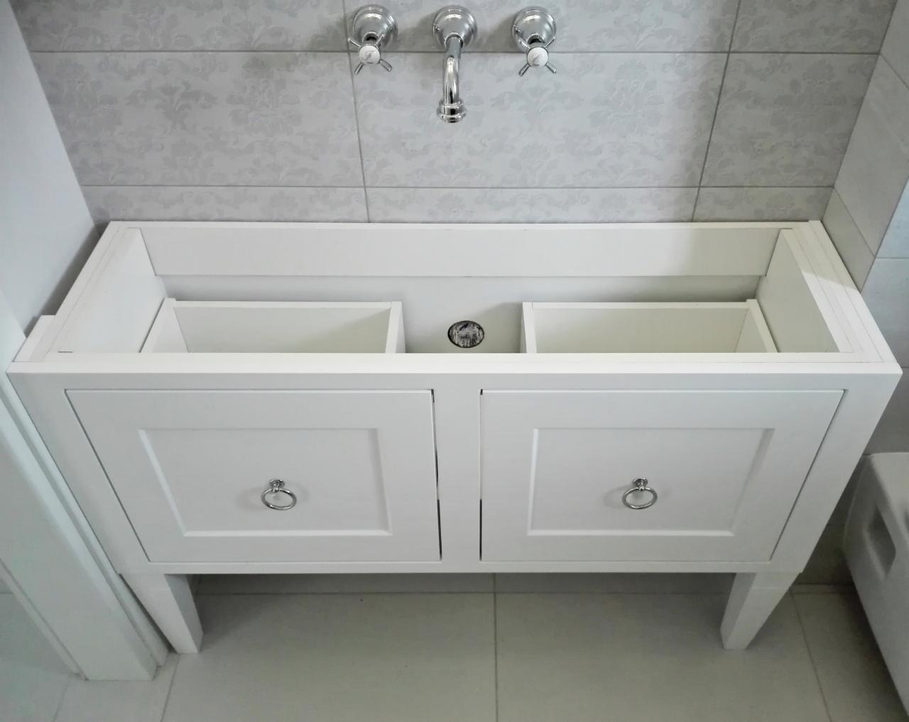 Biała, lakierowana szafka pod umywalkę z dwiema szufladami i srebrnymi uchwytami, bez umywalki, na tle jasnych płytek z delikatnym wzorem.