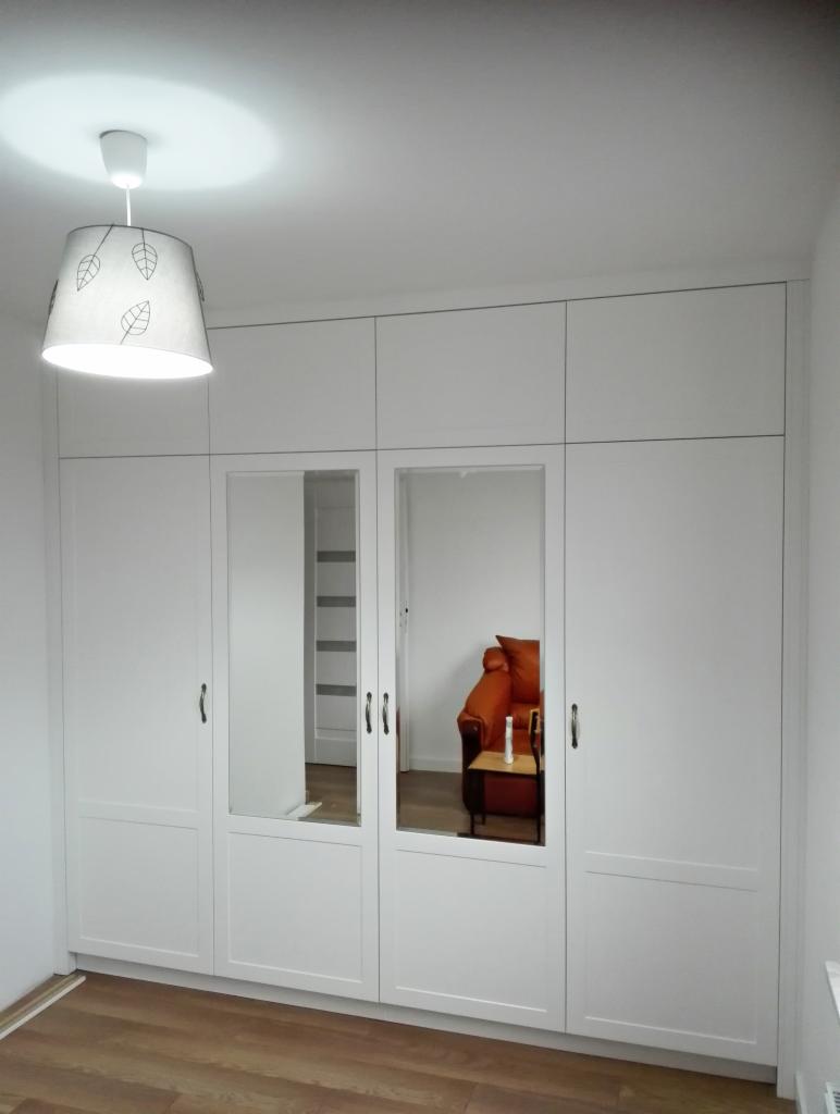 Biała szafa wnękowa z lustrzanymi frontami, odbijająca pomarańczowy fotel i białą drabinkę w tle, oświetlona lampą z motywem liści.