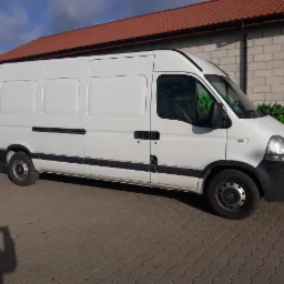 Biały samochód dostawczy Renault Master zaparkowany na brukowanym placu przed budynkiem z czerwoną dachówką, z zielonymi maszynami rolniczymi w tle.