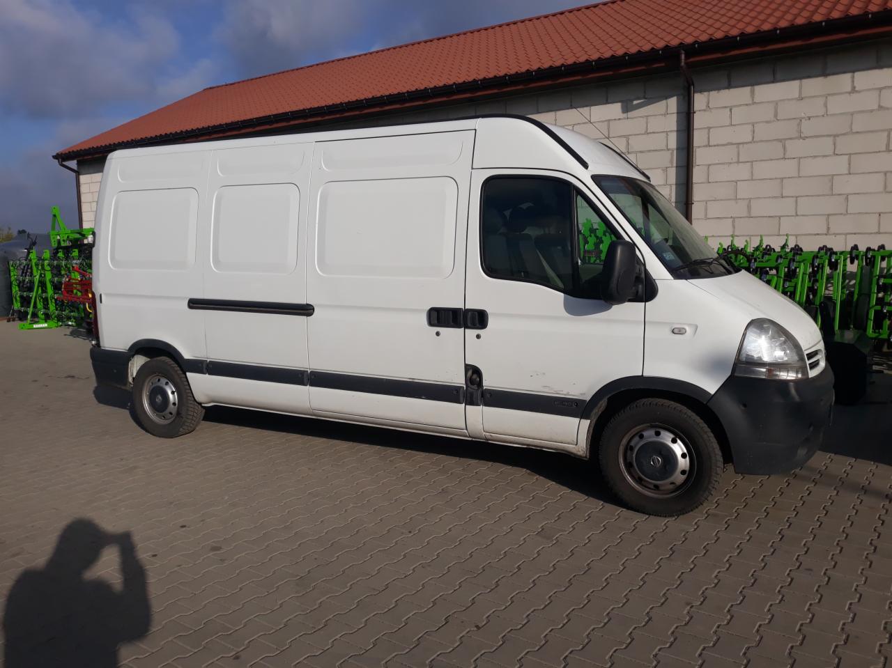 Biały samochód dostawczy Renault Master zaparkowany na brukowanym placu przed budynkiem z czerwoną dachówką, z zielonymi maszynami rolniczymi w tle.